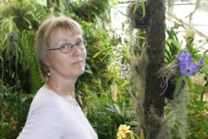 Eine Frau mit blonden kurzen Haaren und einer Brille ist umgeben von großen grünen Pflanzen