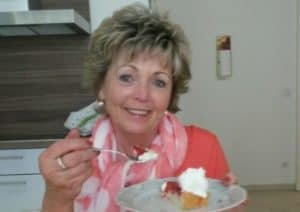 Eine Frau schaut lächelnd in die Kamera und hat in ihrer Hand einen Teller mit Erdbeerkuchen.