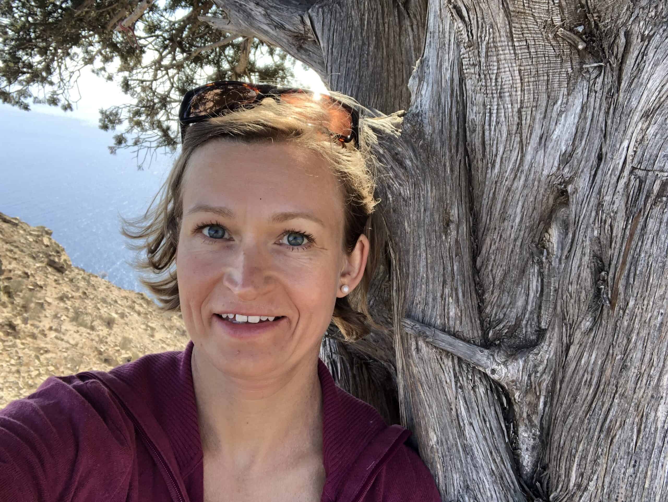 Eine Frau steht an einen Baum gelehnt, lächelt in die Kamera und hinter ihr ist das Meer zu sehen.
